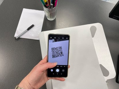 Eine Person hält ein Smartphone in der Hand und scannt einen QR-Code. Auf dem Tisch steht ein Becher mit stiften und ein Block mit Kästchen liegt daneben