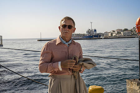 Daniel Craig als Detektiv steht in Hemd und Sonnenbrille an einer Küste.